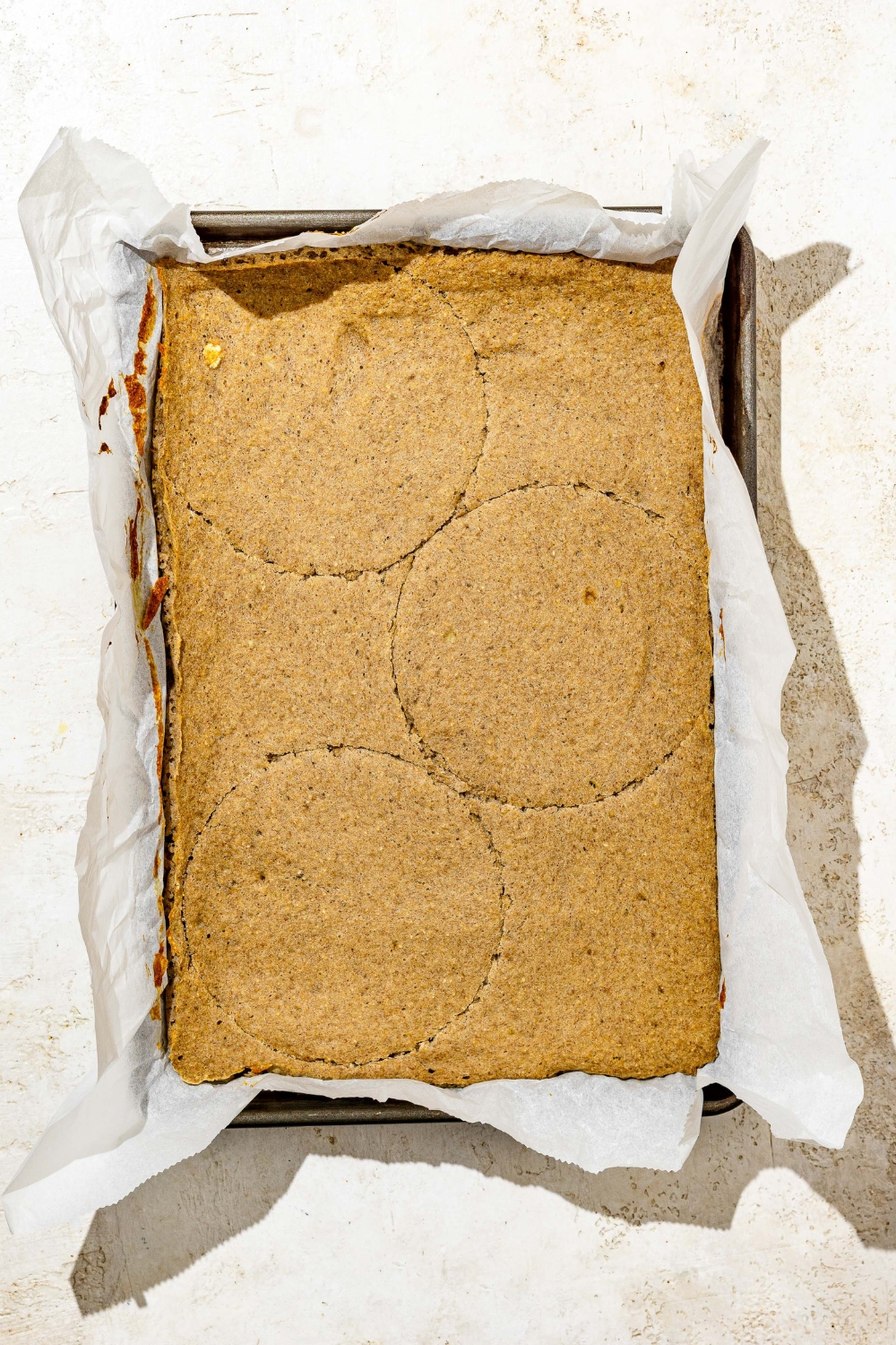 A lined baking pan with baked banana smash cake with three round moulds cut into the cake. The pan is on a tan counter.
