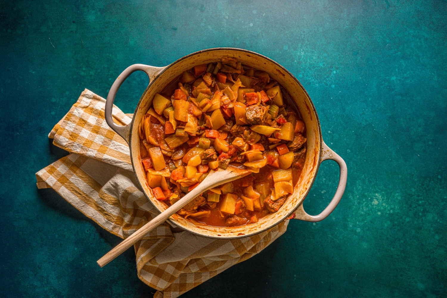 A dutch oven with beef cabbage stew on a blue counter with a striped napkin. A wooden spoon is stirring the stew.