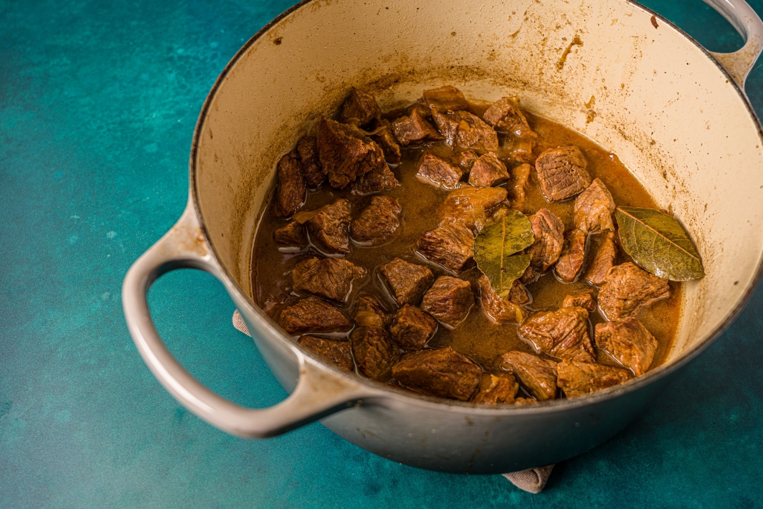 A dutch oven with cubed browned beef cooking in stew sauce with bay leaves. The dutch oven is on a blue counter.