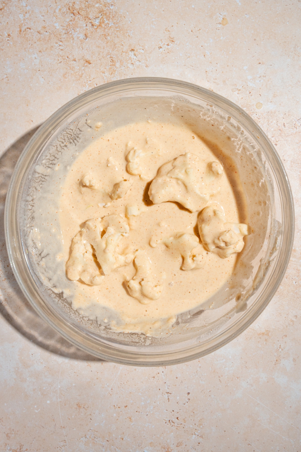 A glass bowl of cauliflower florets submerged in a seasoned batter. The bowl is on a tan counter.