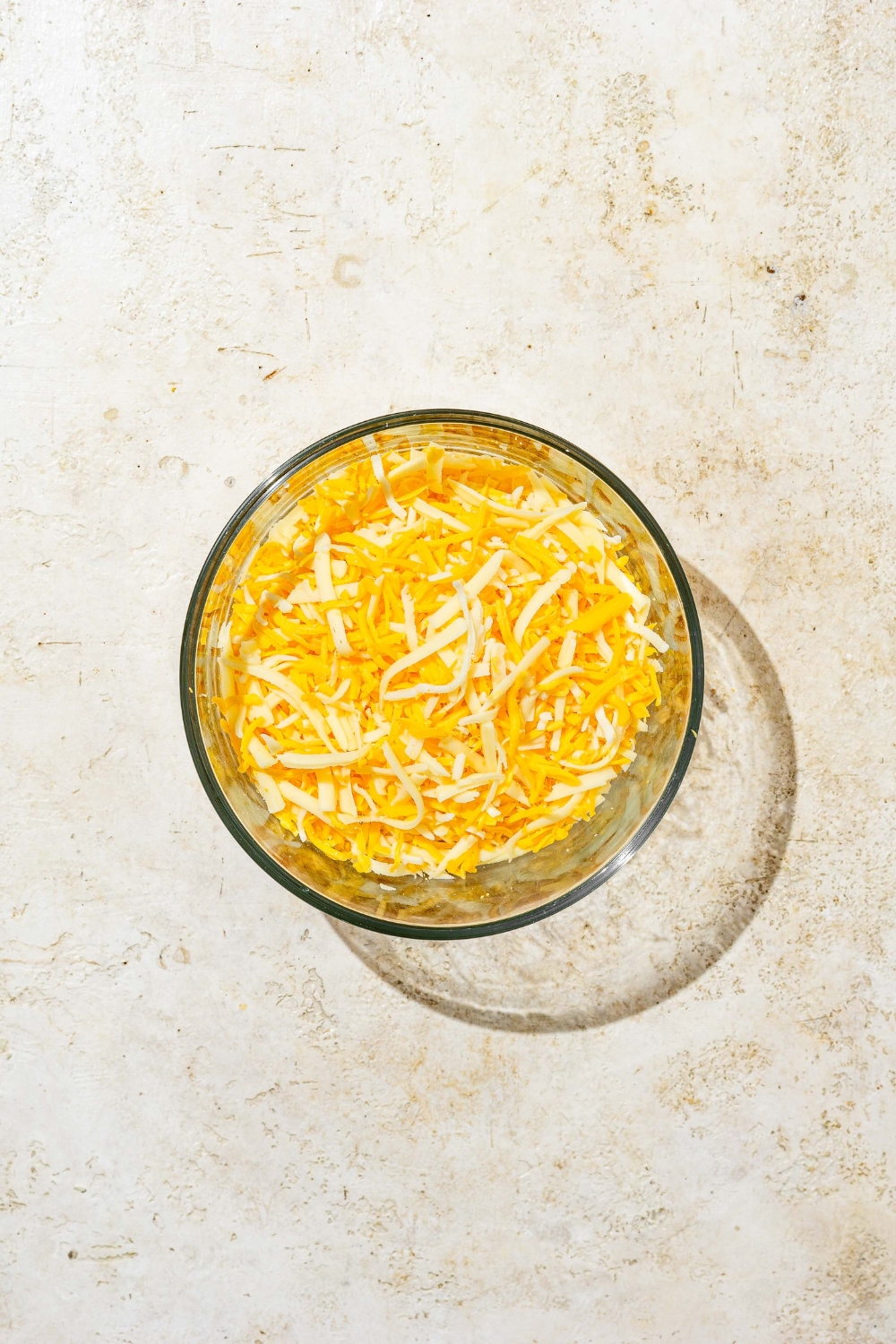A glass bowl of shredded cheeses. The bowl is on a tan counter.