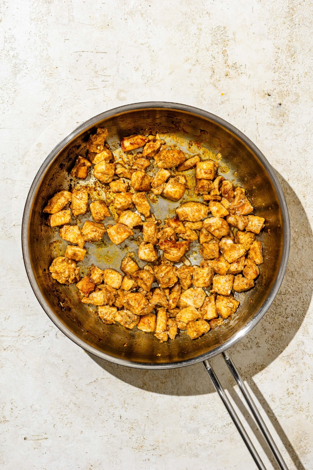 A skillet with cubed seasoned chicken cooking in oil. The skillet is on a tan counter.
