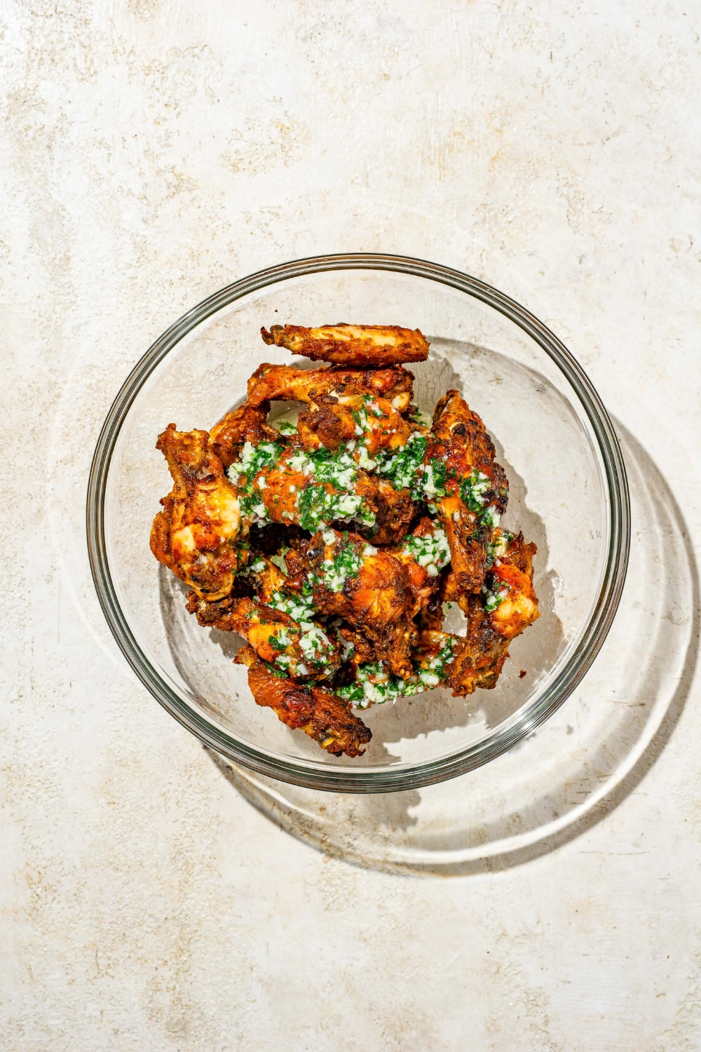 A glass bowl with baked seasoned wings with a parsley butter sauce drizzled on top. The bowl is on a tan counter.