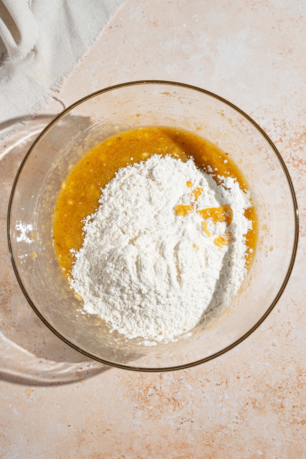 A glass bowl with dry ingredients mixture on top of a wet ingredients mixture for brown butter banana bread. The bowl is on a tan counter.