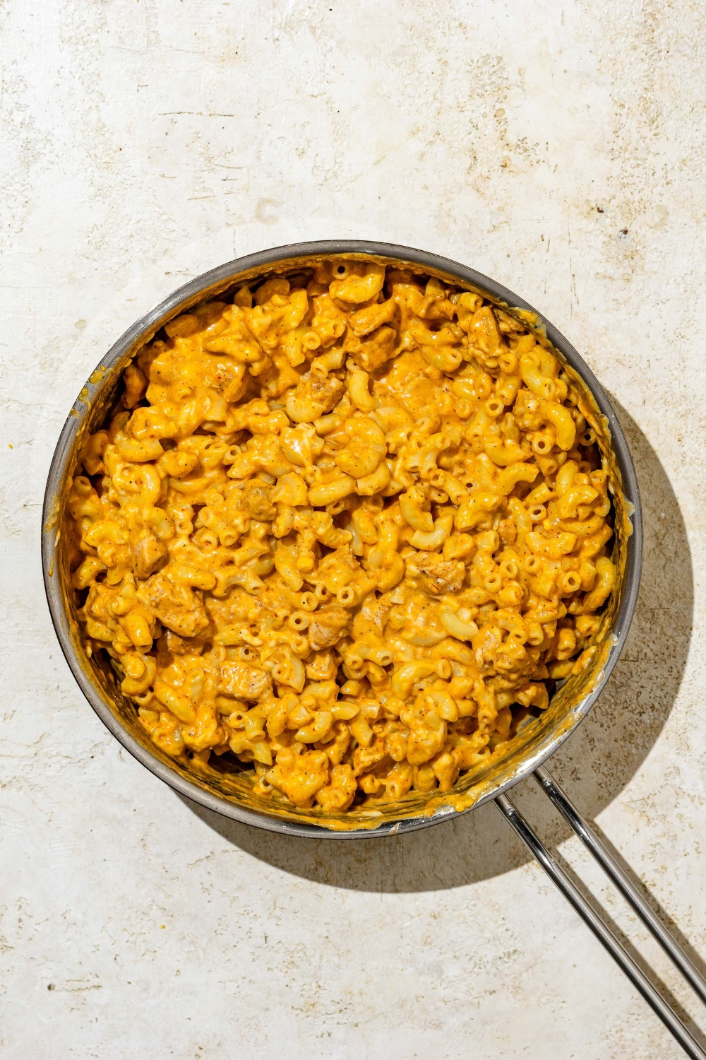 A skillet with elbow pasta mixed with Buffalo mac and cheese sauce. The skillet is on a tan counter.