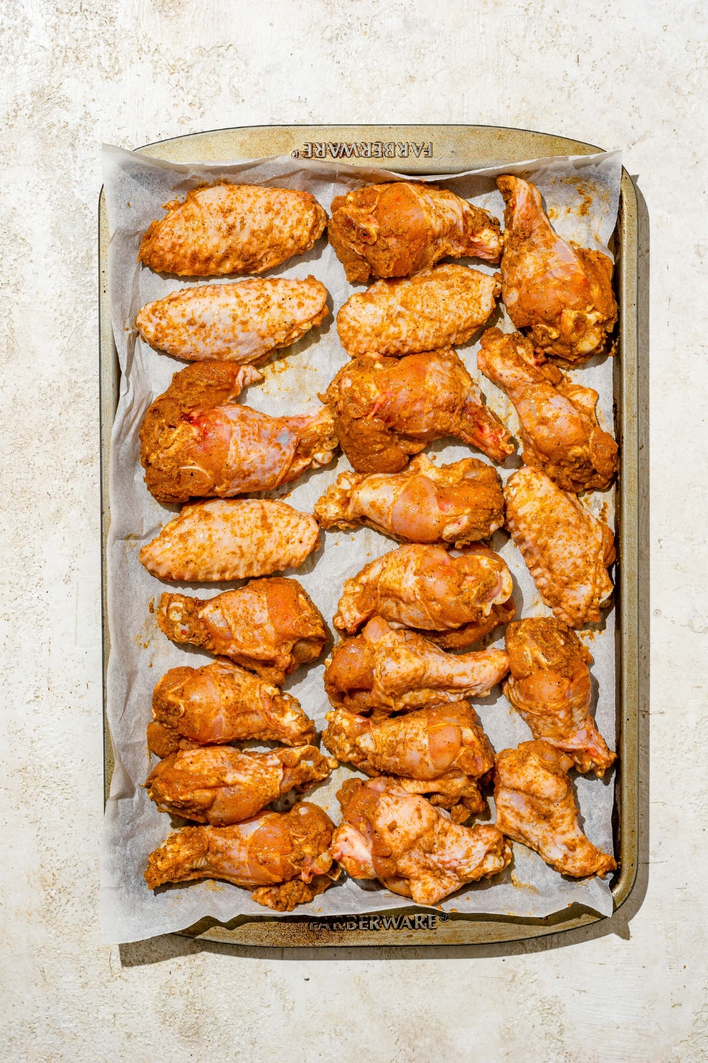 A baking sheet lined with parchment paper with seasoned chicken wings. The sheet is on a tan counter.