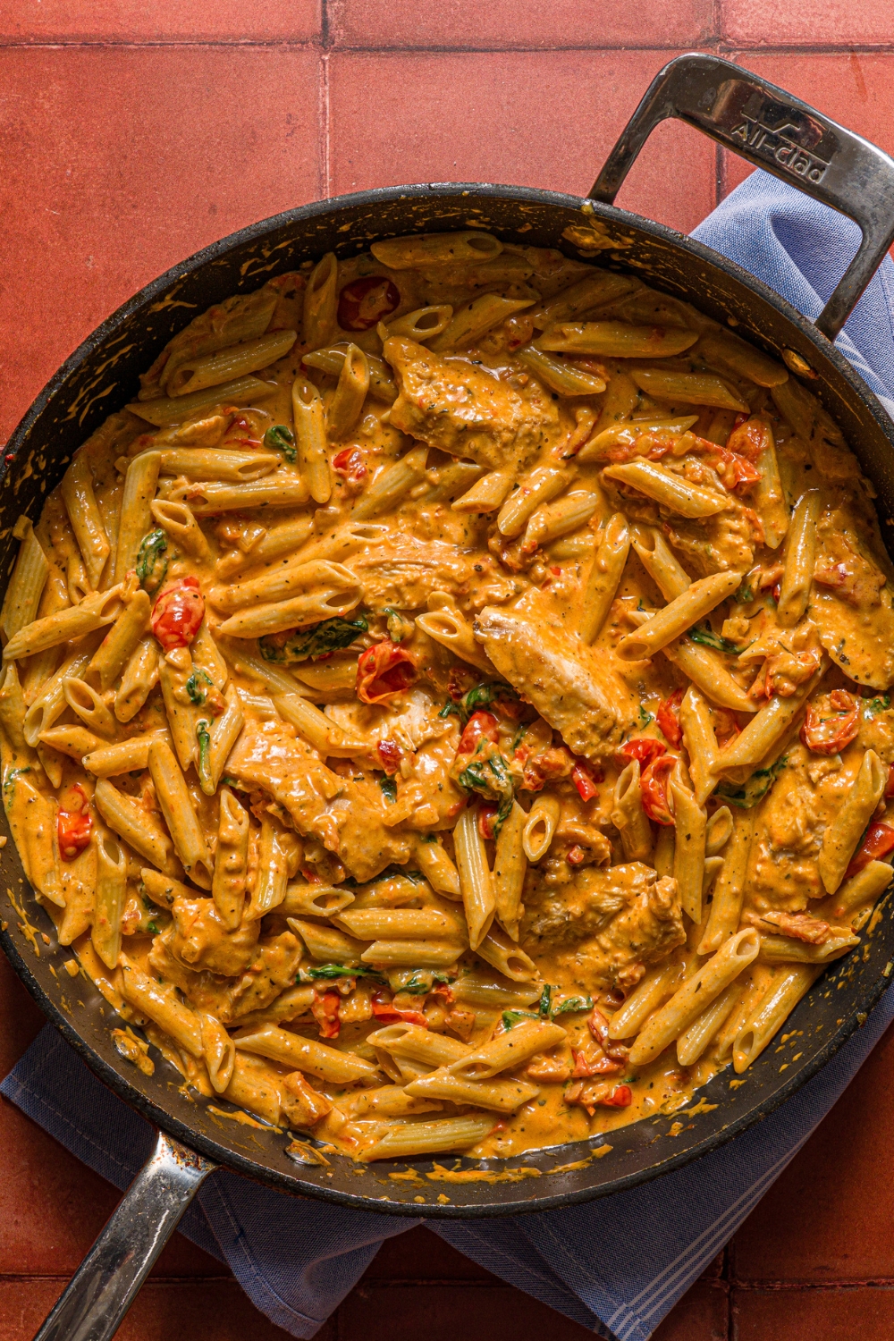 A skillet with tuscan chicken pasta tossed with tomatoes, spinach, and chicken. The skillet is on a tiled counter with a blue cloth napkin.