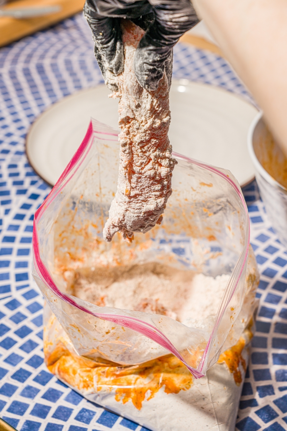 A chicken tender being dipped and dredged into a plastic bag of coating. The bag is on a blue tiled counter.