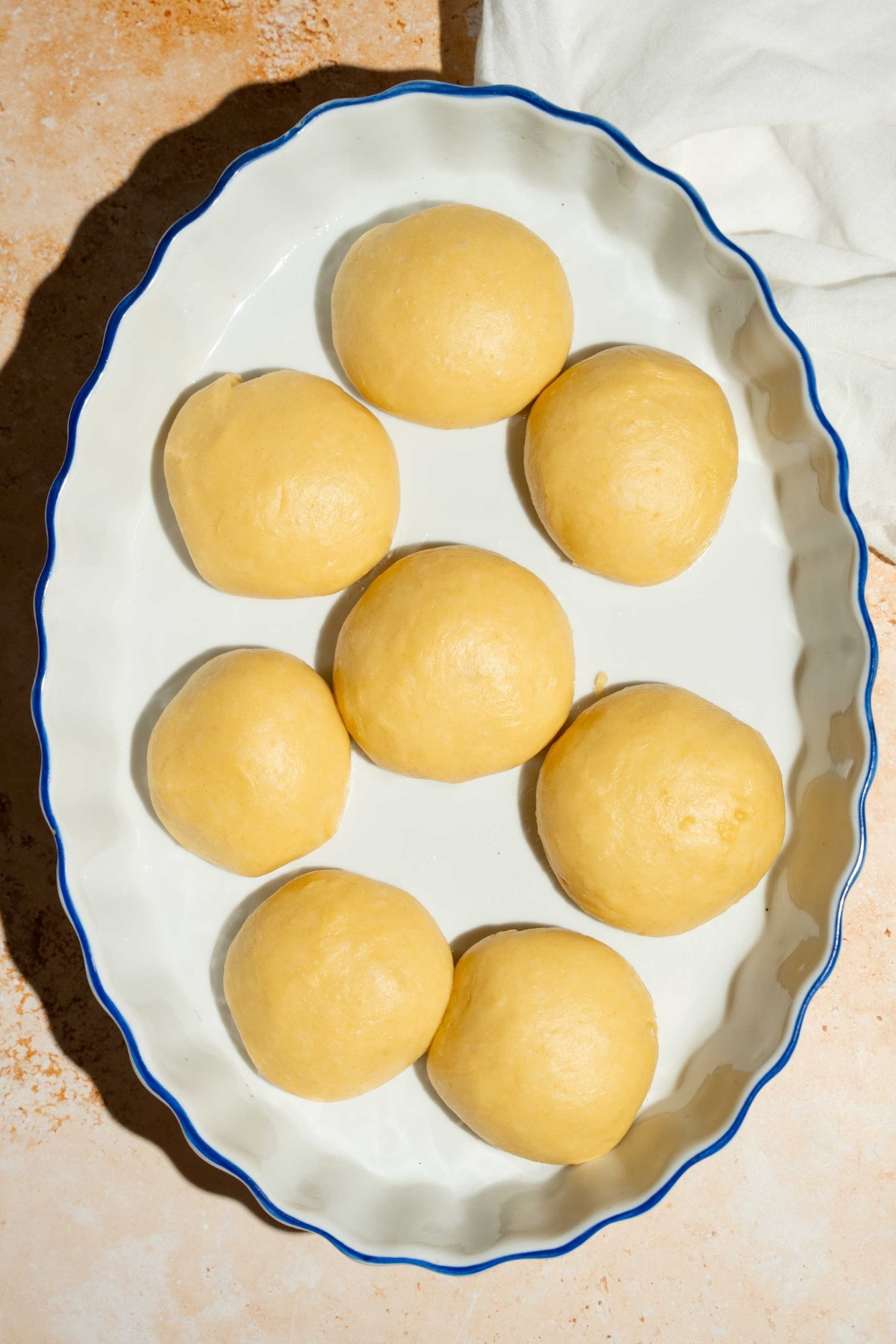 A baking dish with several balls of dinner roll dough. The dish is on a tan counter with a white cloth napkin.