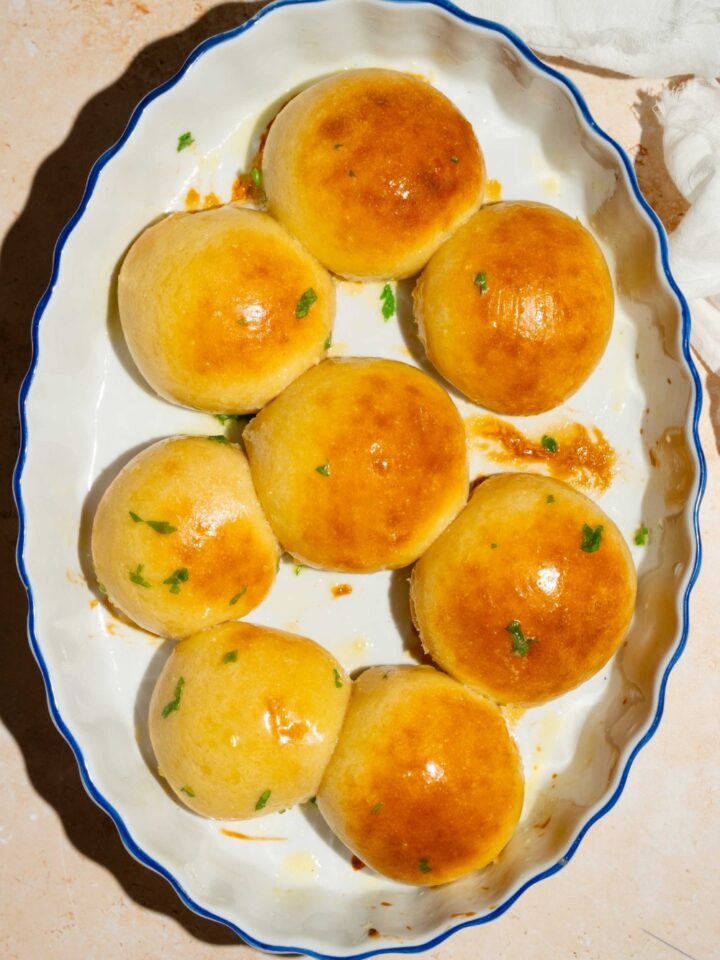 A baking plate with dinner rolls brushed with garlic herb butter and garnished with fresh parsley. The plate is on a tan counter with a white cloth napkin.