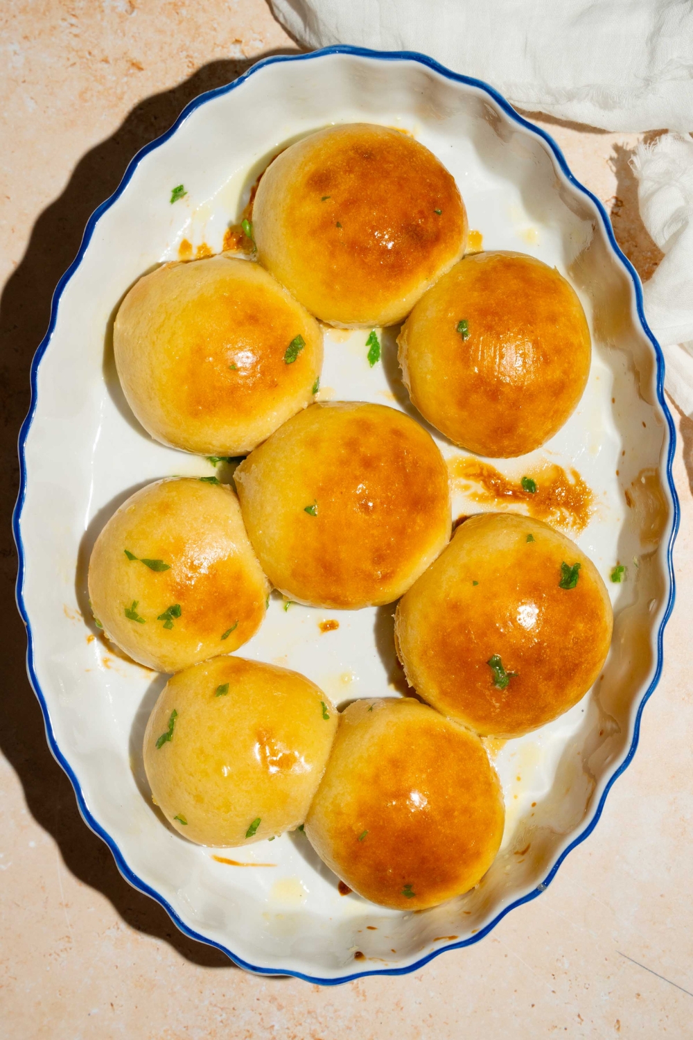 A baking plate with dinner rolls brushed with garlic herb butter and garnished with fresh parsley. The plate is on a tan counter with a white cloth napkin.