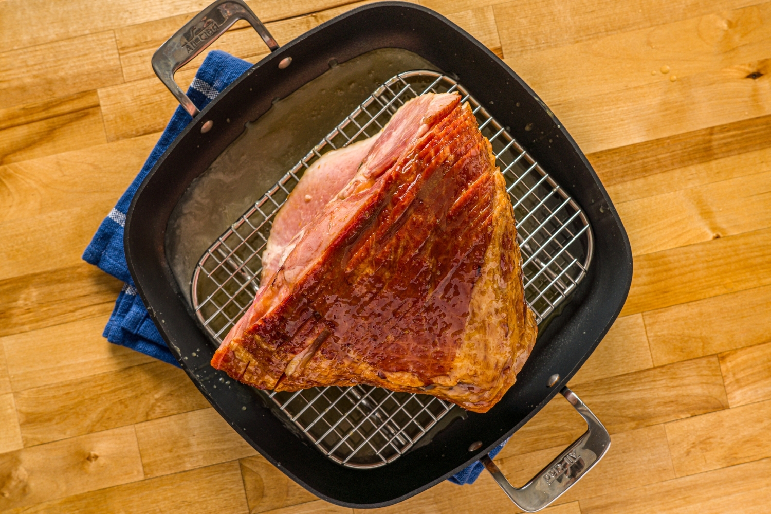 A glazed spiral ham on a roasting pan with a rack inside the pan. The pan is on a wooden counter with a blue cloth napkin.