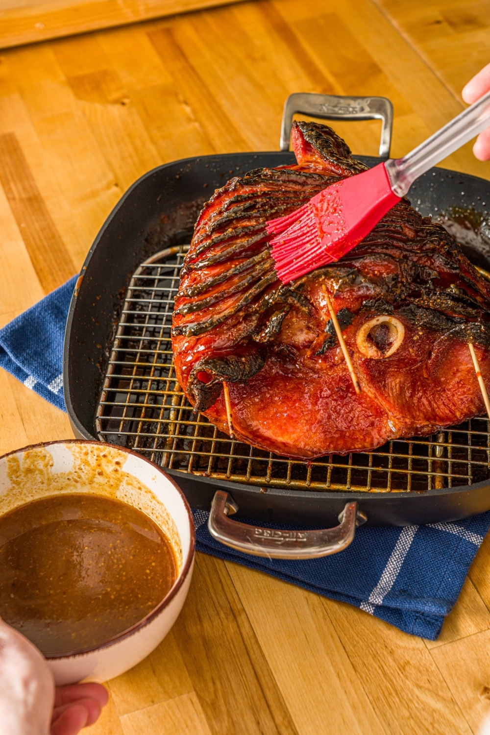 A glazed spiral ham on a roasting pan lined with a rack. The ham is being brushed with glaze. The pan is on a wooden counter with a blue cloth napkin and small bowl of glaze.