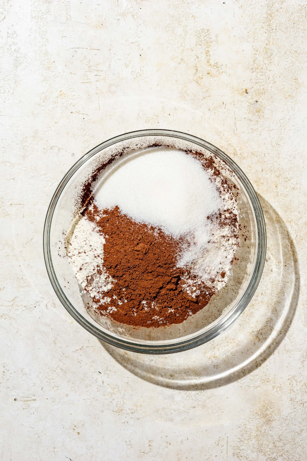 A glass bowl with dry ingredients to make chocolate cinnamon roll dough including four, cocoa powder, and sugar. The bowl is on a tan counter.