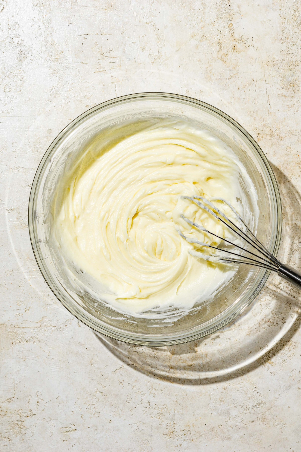 A white bowl with a whisk mixing ingredients to make white Texas sheet cake icing including powdered sugar, butter, sour cream, and vanilla. The bowl is on a white counter.