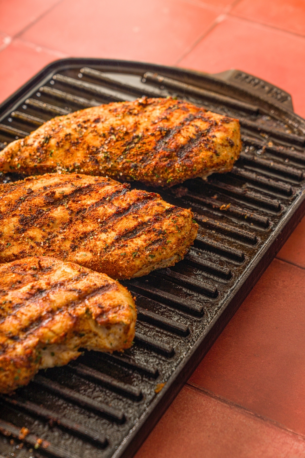 Three seasoned chicken breasts grilling on an indoor grill. The grill is on a red counter.