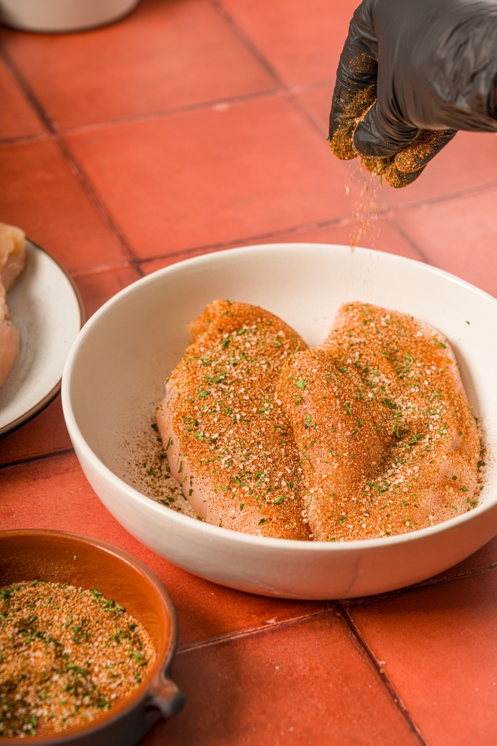 A bowl of uncooked chicken breasts tossed in seasonings. There is a hand sprinkling seasonings over the chicken. The bowl is on a red counter.