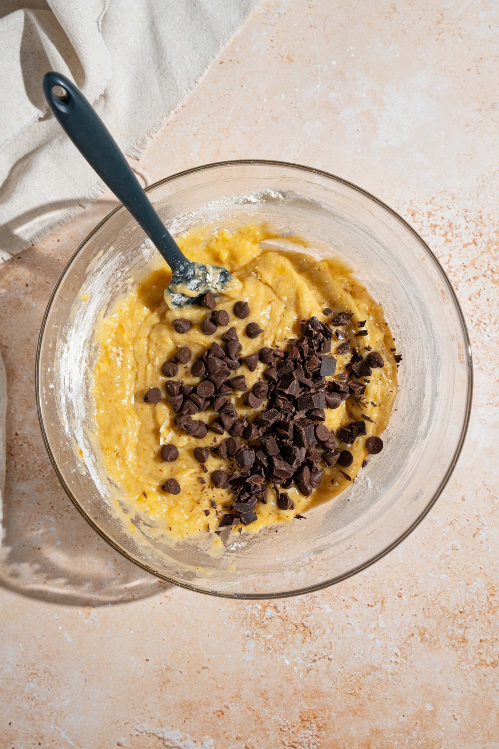 A glass bowl with a spatula folding in chocolate chips into a brown butter banana bread batter. The bowl is on a tan counter.
