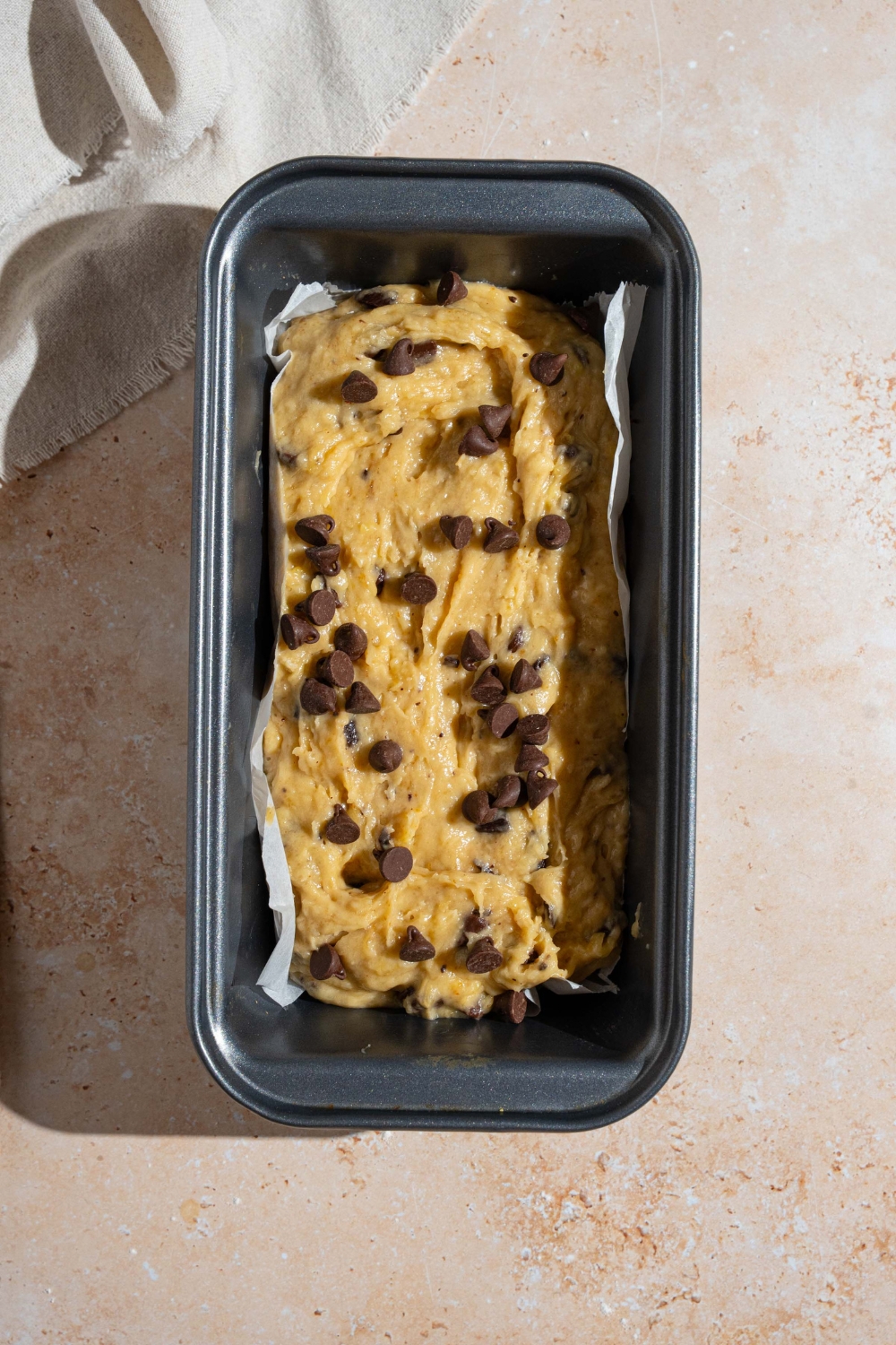 A bread pan lined with parchment paper with brown butter chocolate chip banana bread batter. The pan is on a tan counter.