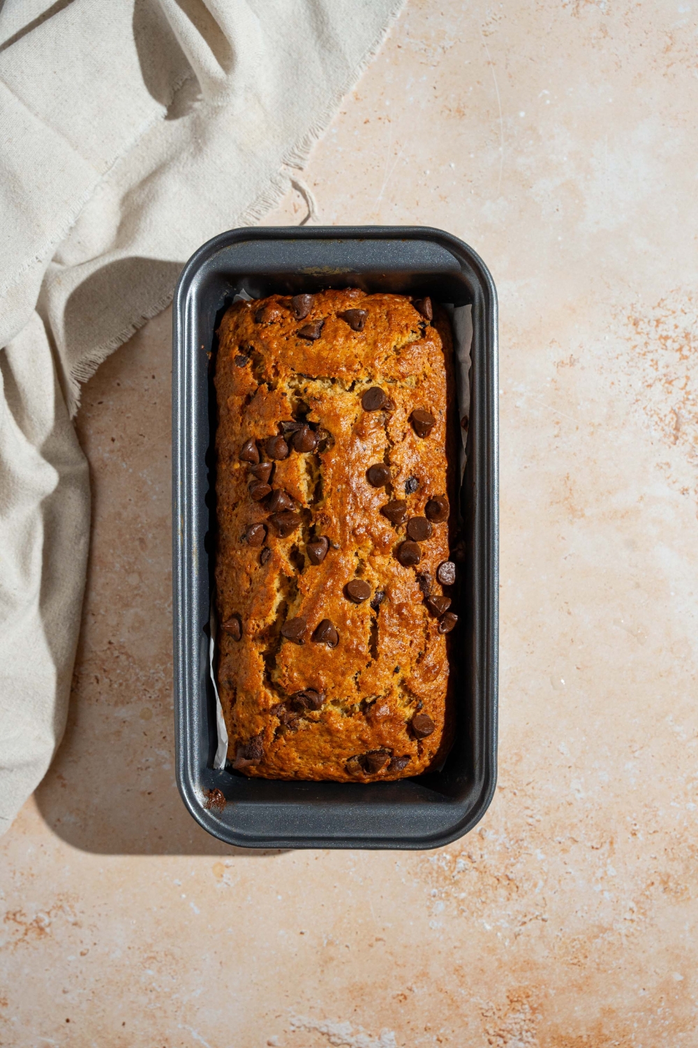 A bread pan with baked brown butter banana bread with chocolate chips. The pan is on a tan counter.