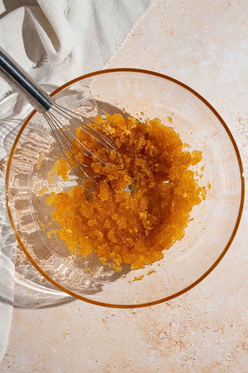 A glass bowl with a whisk combining brown sugar and brown butter. The bowl is on a tan counter.