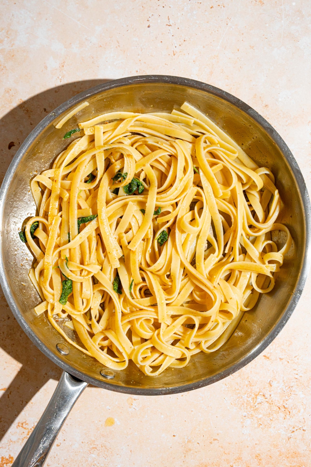 A skillet with linguine pasta tossed in brown butter sauce and sage. The skillet is on a tan counter.
