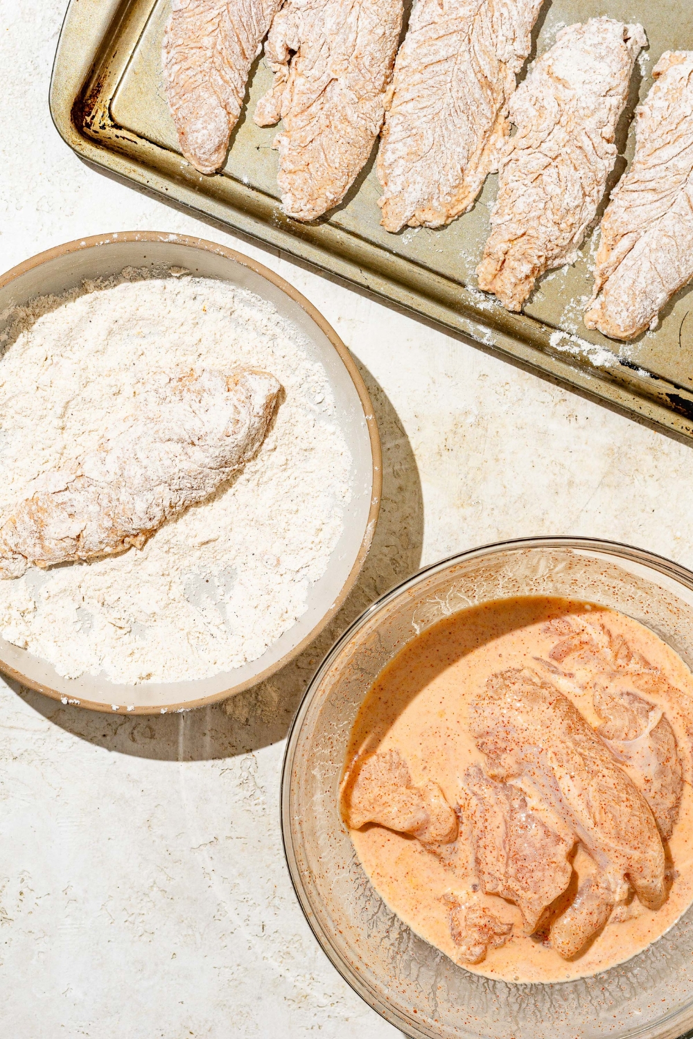 An uncooked chicken tender coated in a bowl of breading. A second bowl of uncooked chicken tenders sitting in a bowl of marinade. And a baking sheet with breaded chicken tenders lined on the sheet.