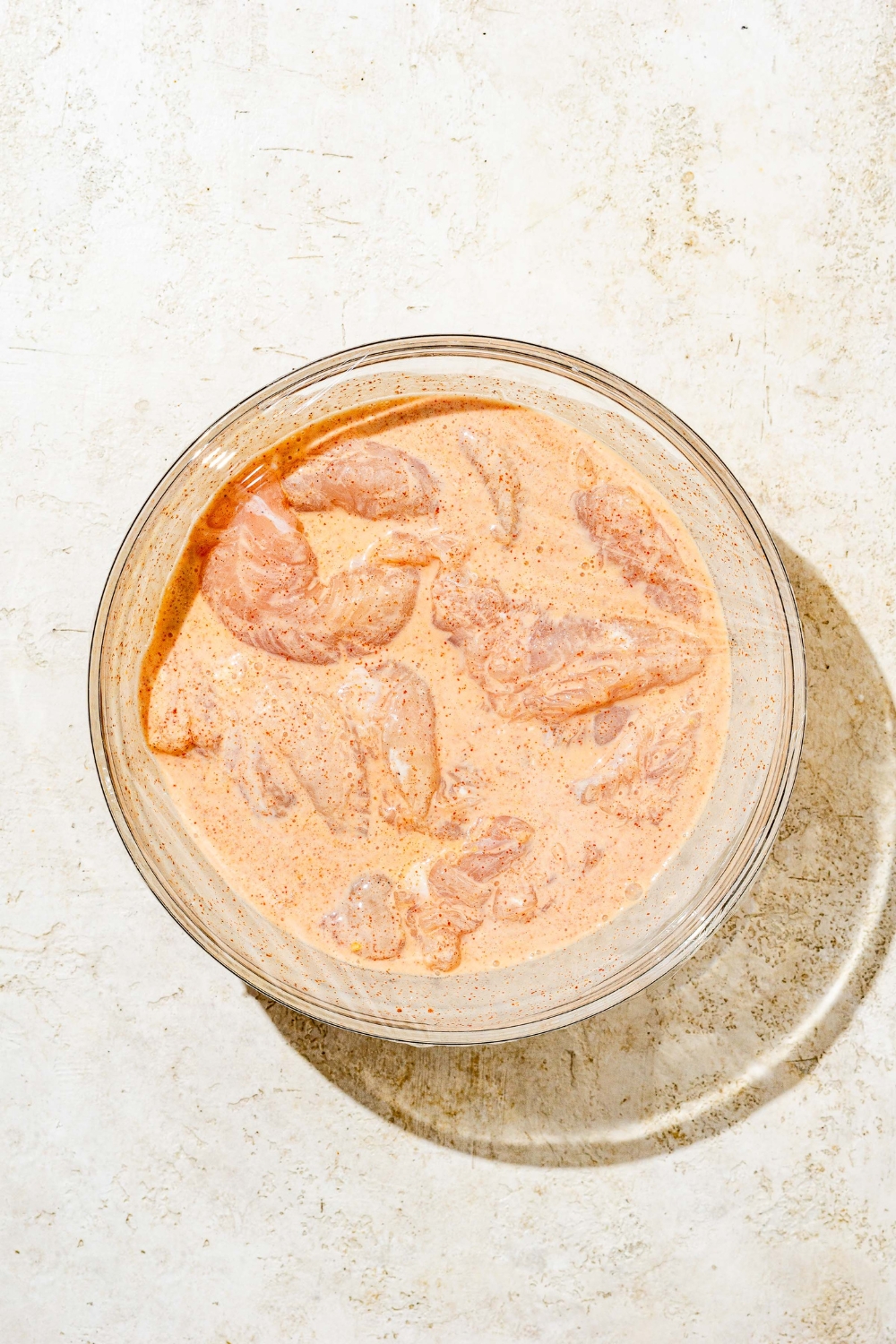 A glass bowl with uncooked chicken tenders sitting in marinade. The bowl is on a white counter.