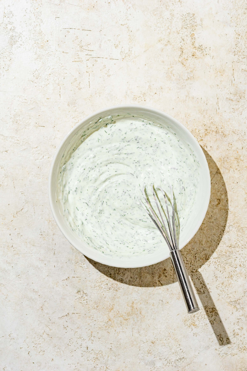 A white bowl with a whisk mixing ingredients to make homemade ranch dressing including buttermilk, sour cream, dill, lemon, and mayo. The bowl is on a white counter.