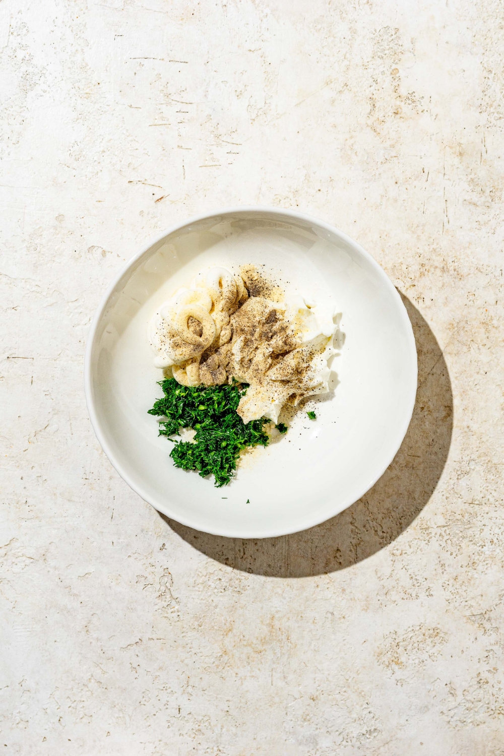 A glass bowl with ingredients to make homemade ranch dressing including mayo, sour cream, dill, and seasonings. The bowl is on a white counter.