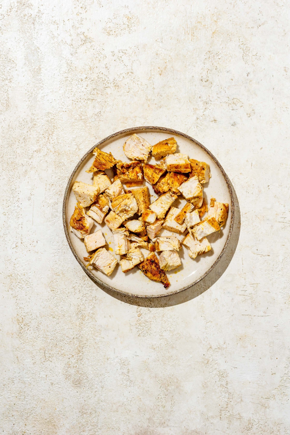 A plate with cubed cooked chicken. The plate is on a tan counter.