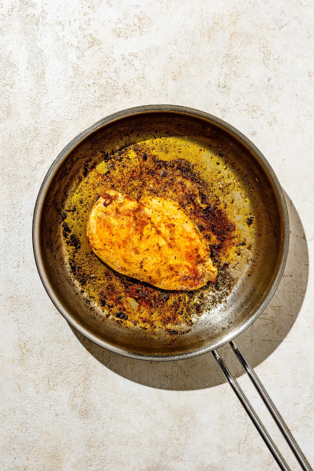 A skillet with a chicken breast cooked in oil and butter. The skillet is on a tan counter.