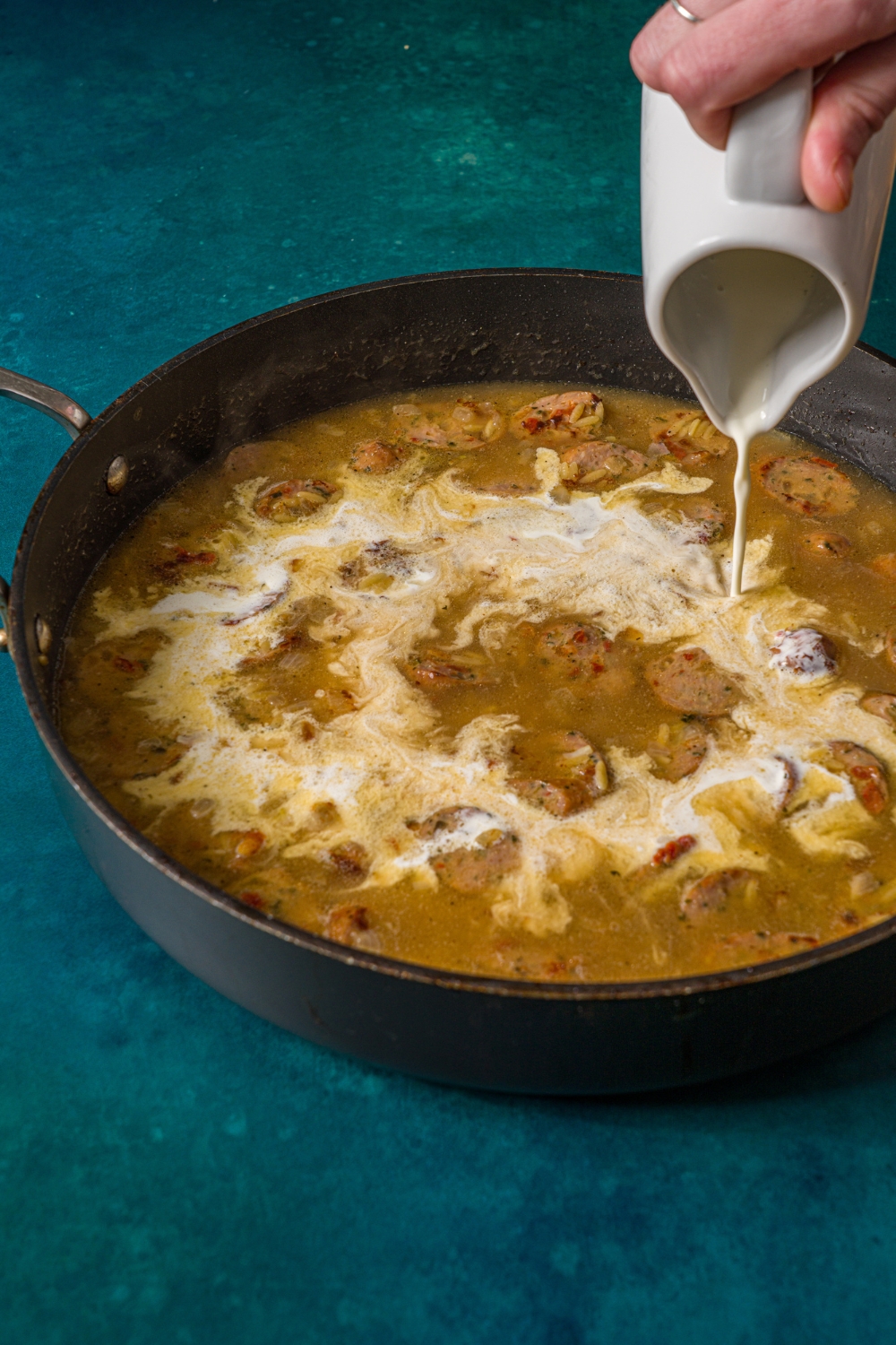A skillet with chicken sausage orzo cooking in chicken broth and seasonings. A hand is pouring heavy cream from a ceramic creamer into the skillet. The skillet is on a blue counter.