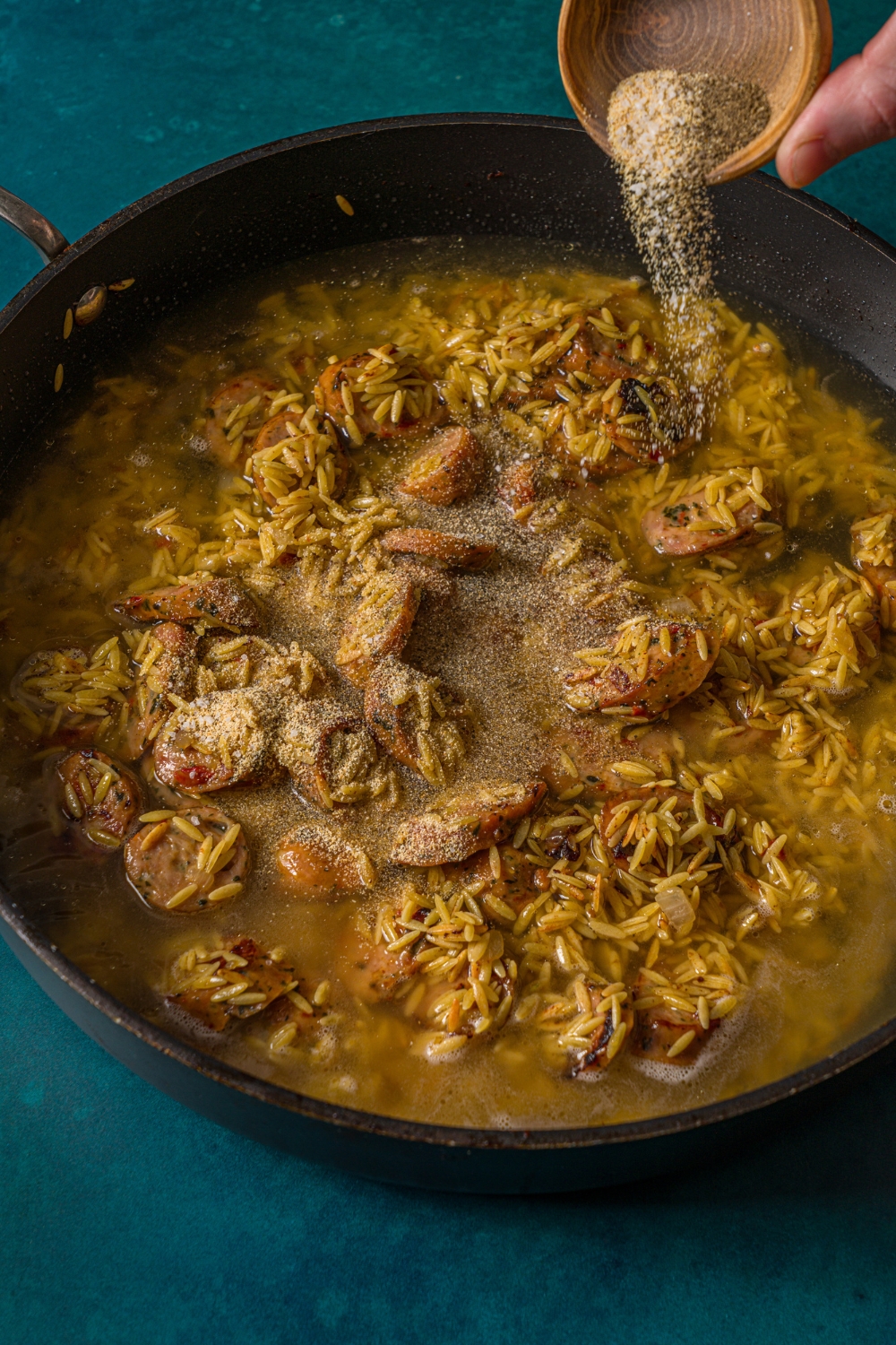 A skillet with chicken orzo cooking in chicken broth. There is a hand pouring a bowl of seasonings into the skillet. The skillet is on a blue counter.