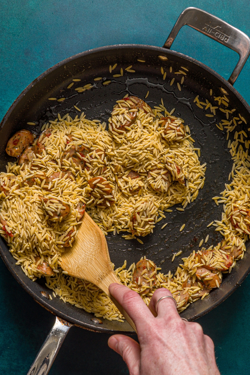 A skillet with sautéed chicken sausage mixed with toasted orzo. A hand is using a wooden spoon to stir the mixture. The skillet is on a blue counter.
