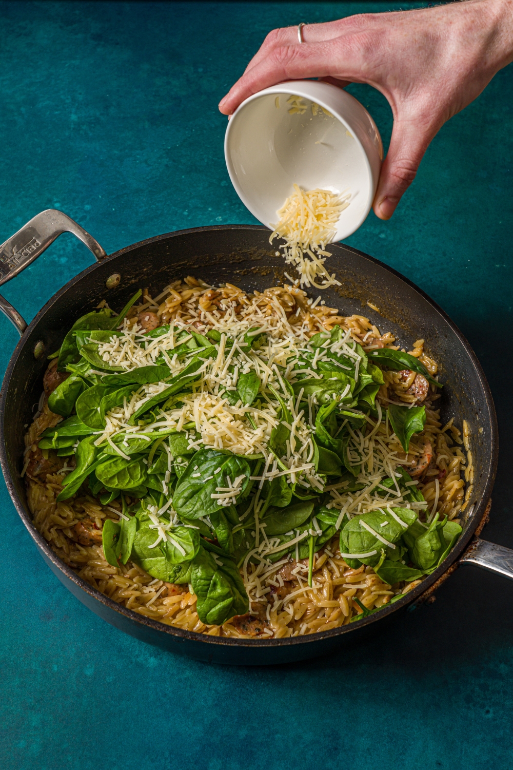 A skillet with chicken sausage orzo cooking with fresh spinach. A hand is pouring. bowl of shredded cheese into the skillet. The skillet is on a blue counter.