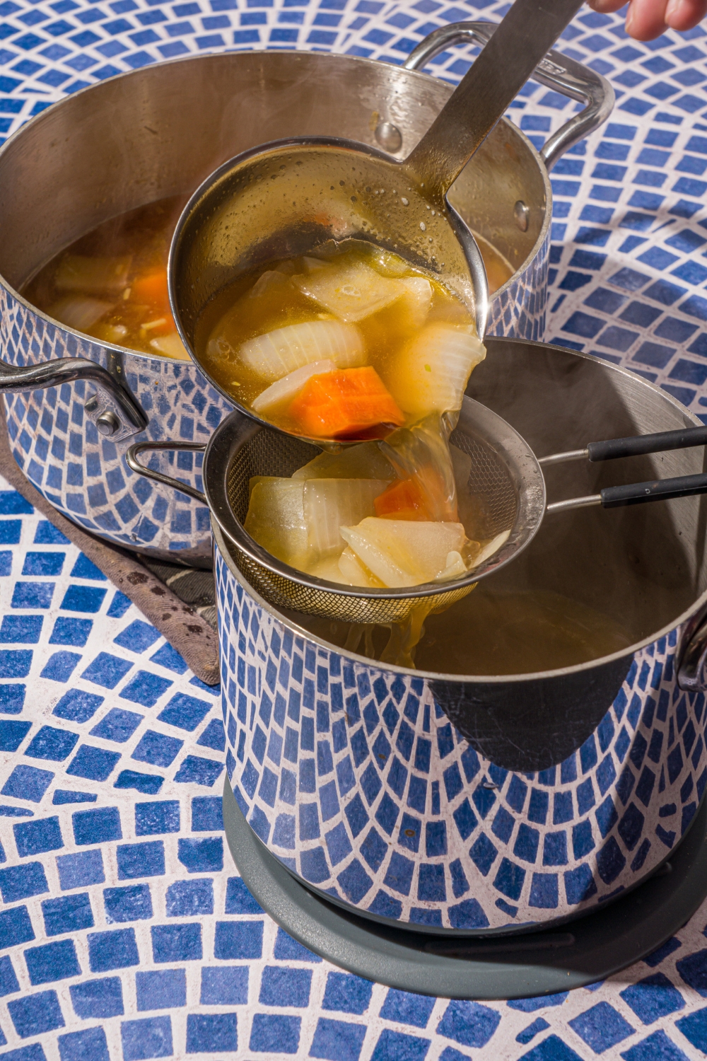 A ladle straining clear soup broth with chopped vegetables through a sieve into another stock pot. The pots are on a blue tiled counter.