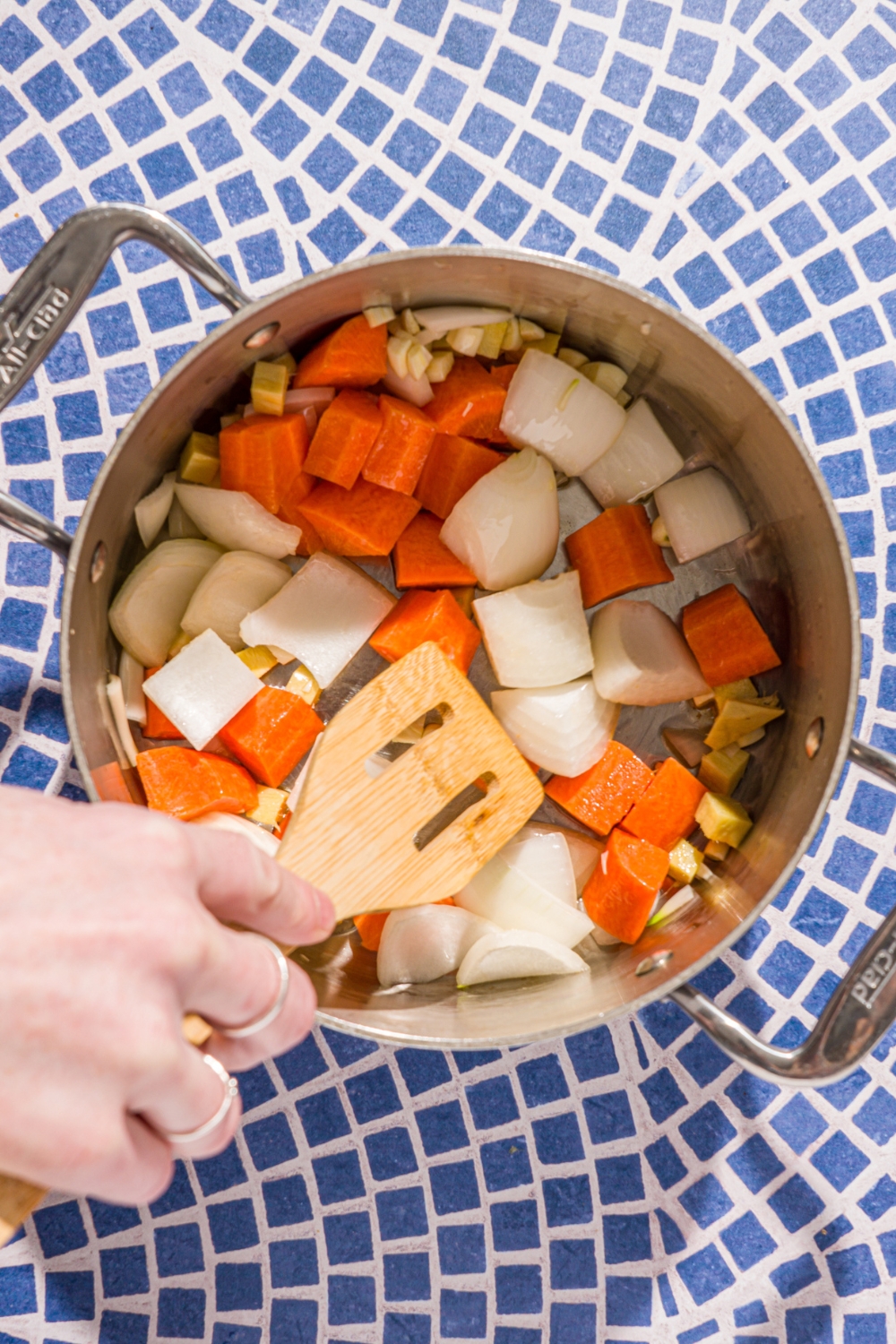 A stock pot with chopped carrots, white onion, ginger, and garlic sautéing in oil. The pot is on a blue tiled counter.