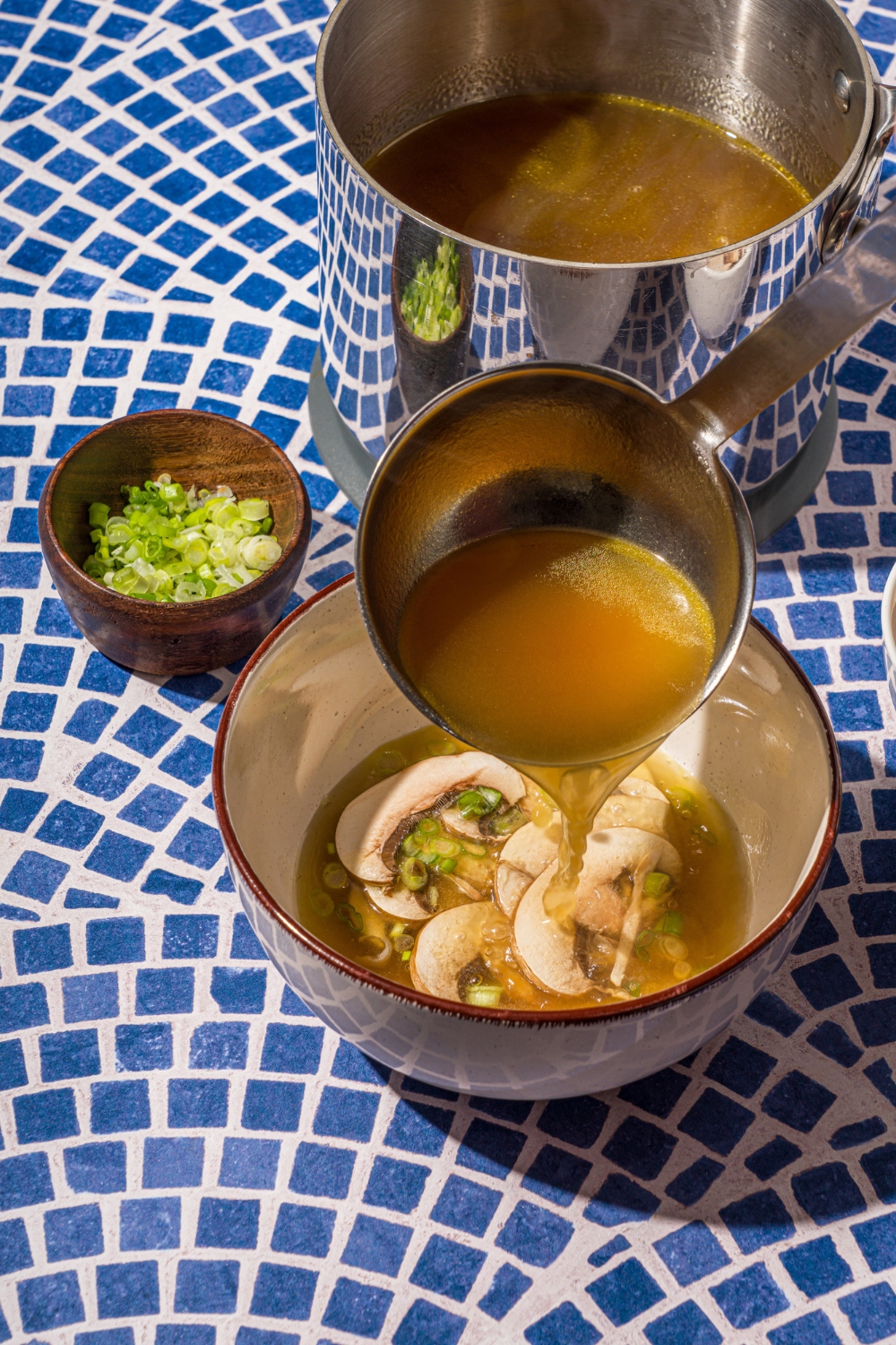 A ceramic bowl with sliced mushrooms and sliced green onions with a ladle pouring broth into the soup. The bowl is on a blue tiled counter with a stock pot of broth and small bowl of sliced green onions.