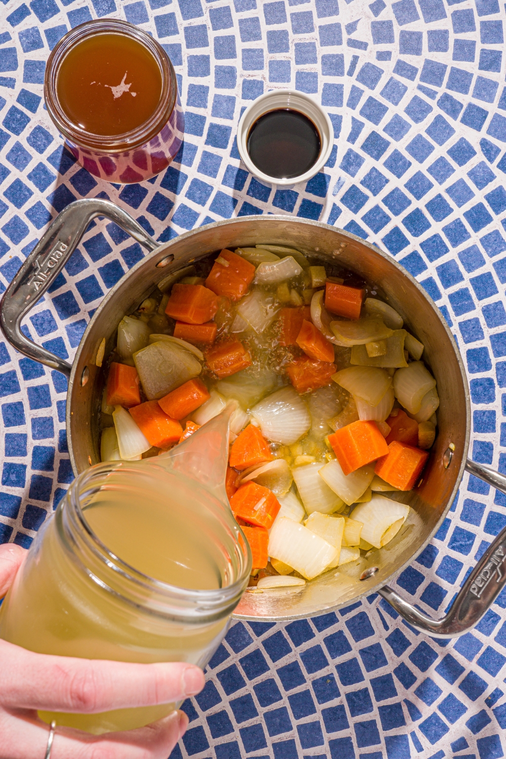 A stock pot with sautéed chopped vegetables. A hand is pouring chicken broth from a jar into the pot. The pot is on a blue tiled counter with a glass jar of beef broth and small bowl of liquid aminos.