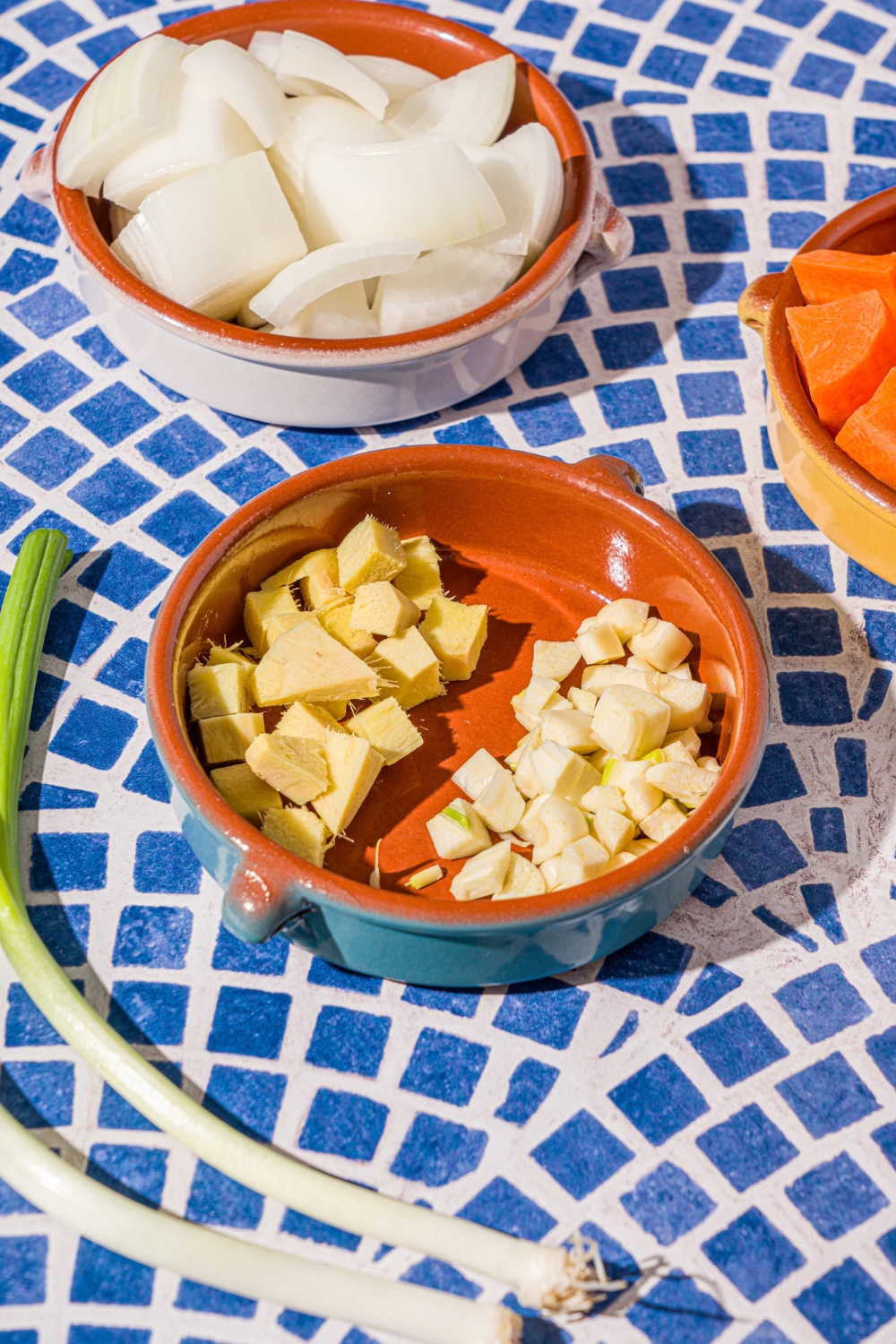 Three ceramic bowls with chopped vegetables including chopped white onion, carrots, ginger, and garlic. The bowls are on a blue tiled counter.