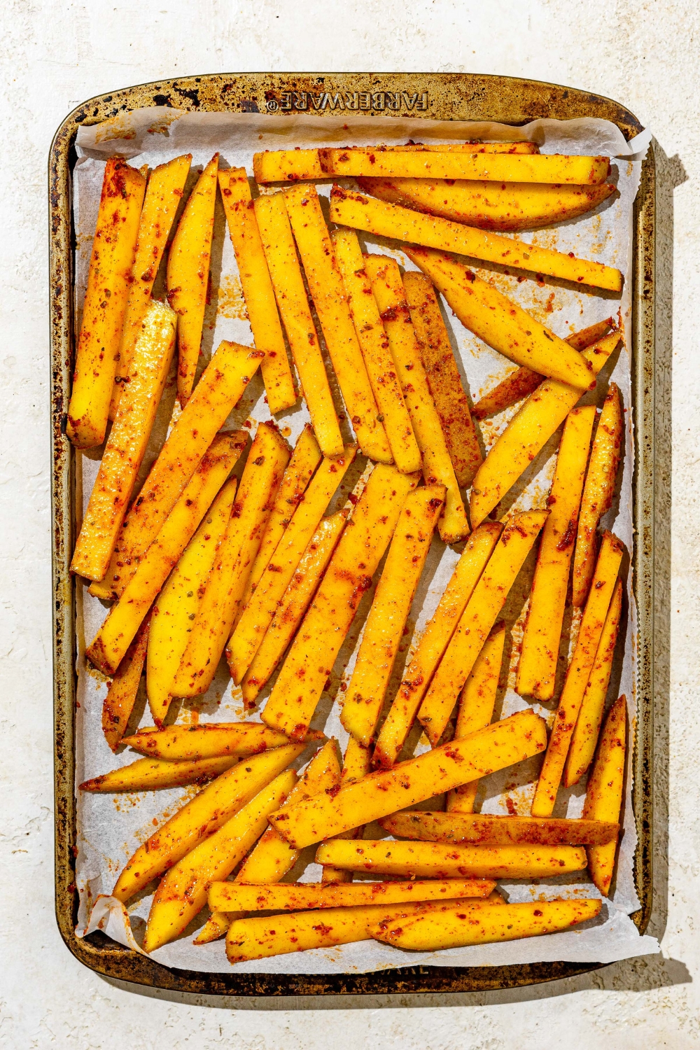A baking sheet lined with parchment paper with uncooked fries tossed in cajun seasoning. The sheet is on a white counter.