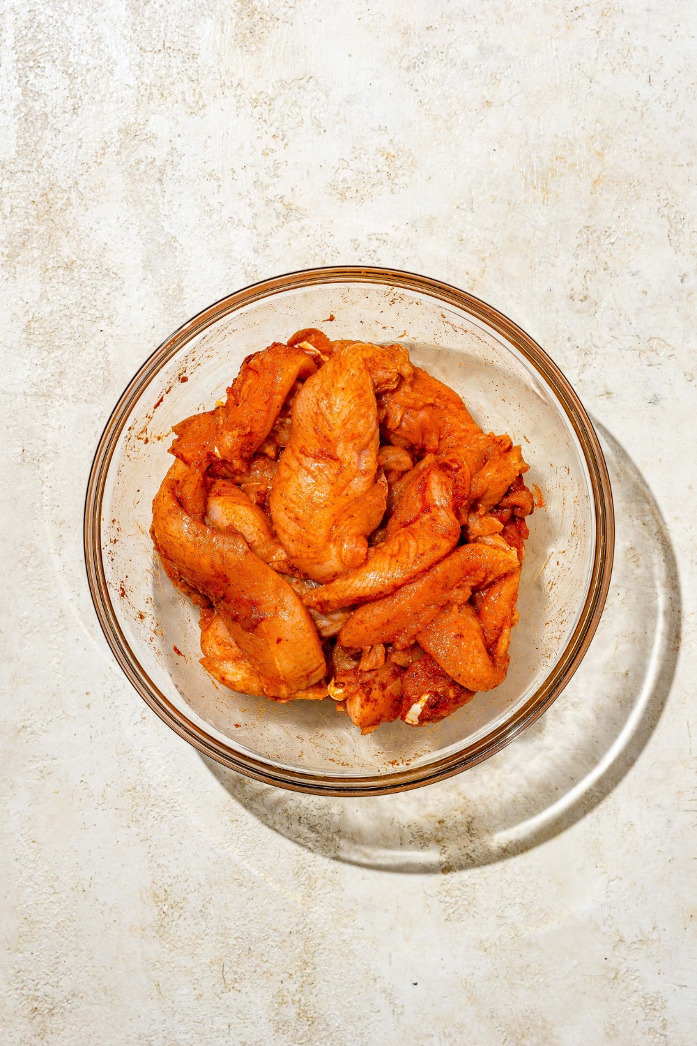A glass bowl with uncooked chicken tenders tossed in seasonings. The bowl is on a tan counter.