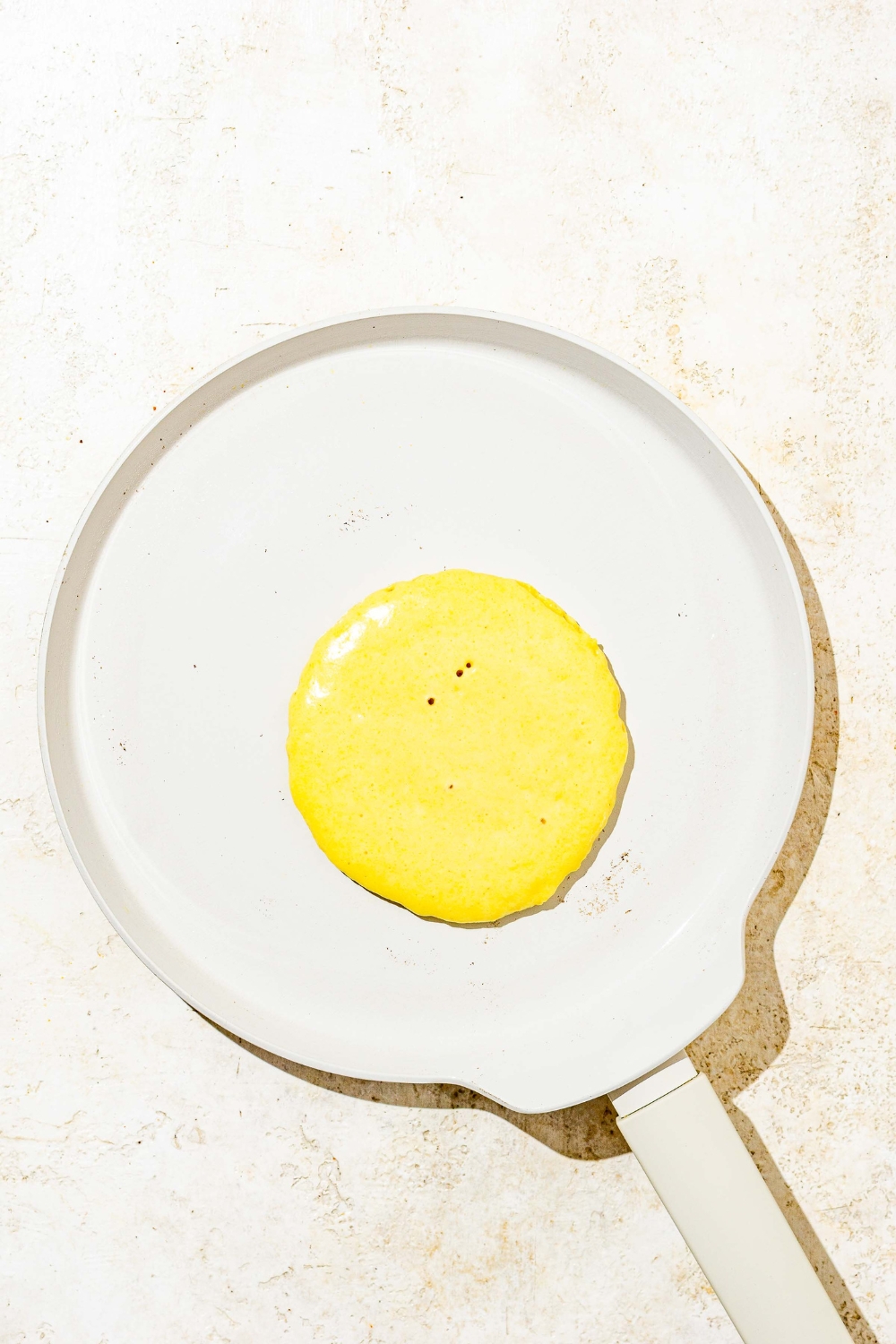 A skillet with an cornmeal pancake cooking on one side. The skillet is on a white counter.