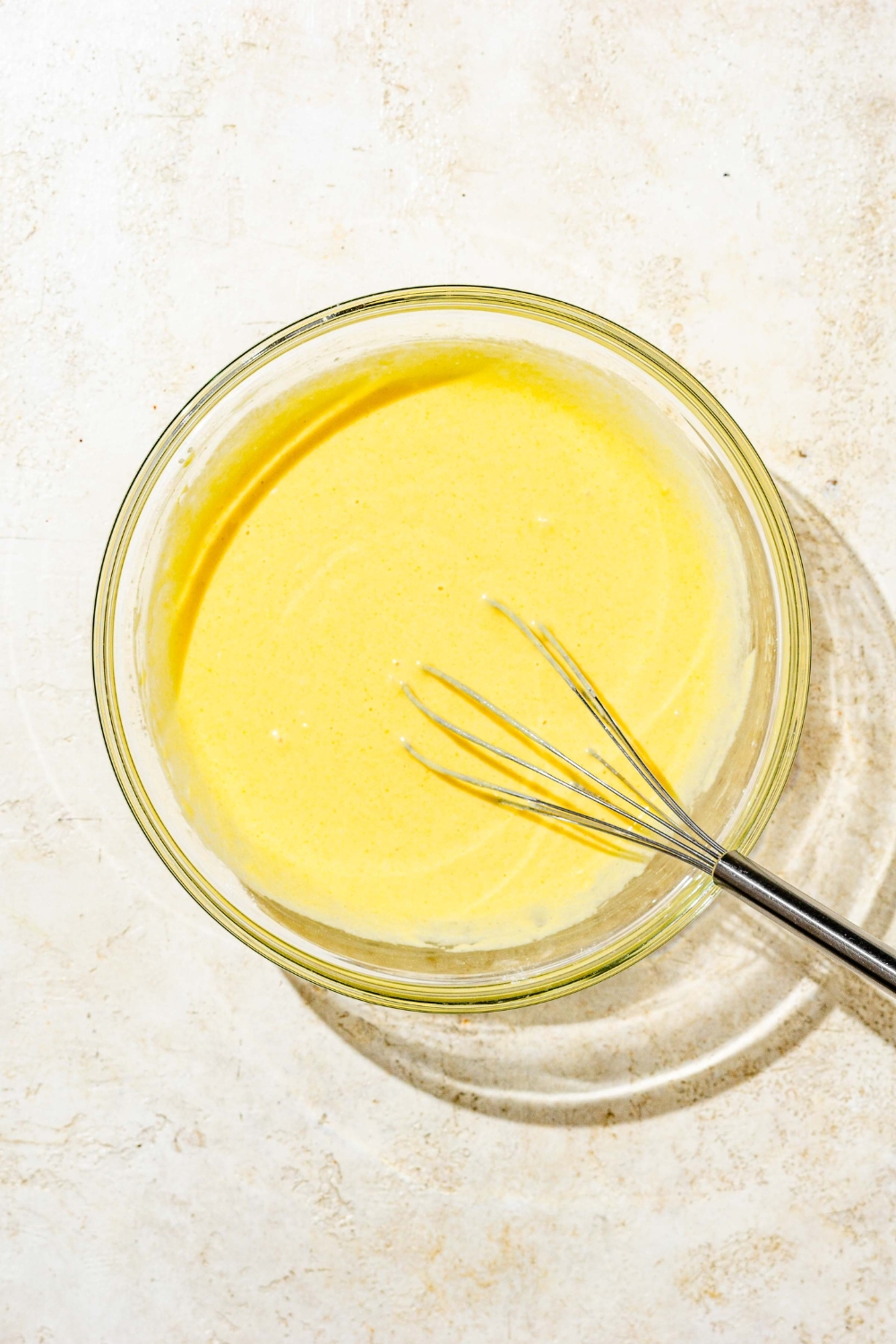 A glass bowl with a whisk mixing cornmeal pancake batter. The bowl is on a white counter.