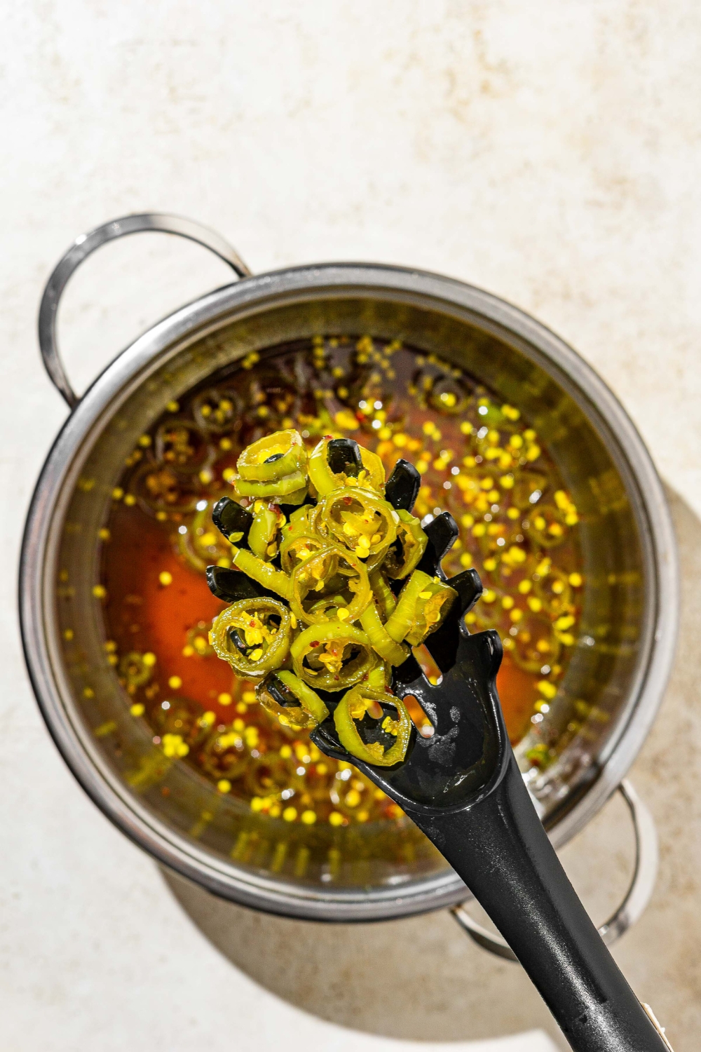A stock pot with cooked candied jalapeños sauce with a slotted spoon of cooked jalapeños. The pot is on a tan counter.