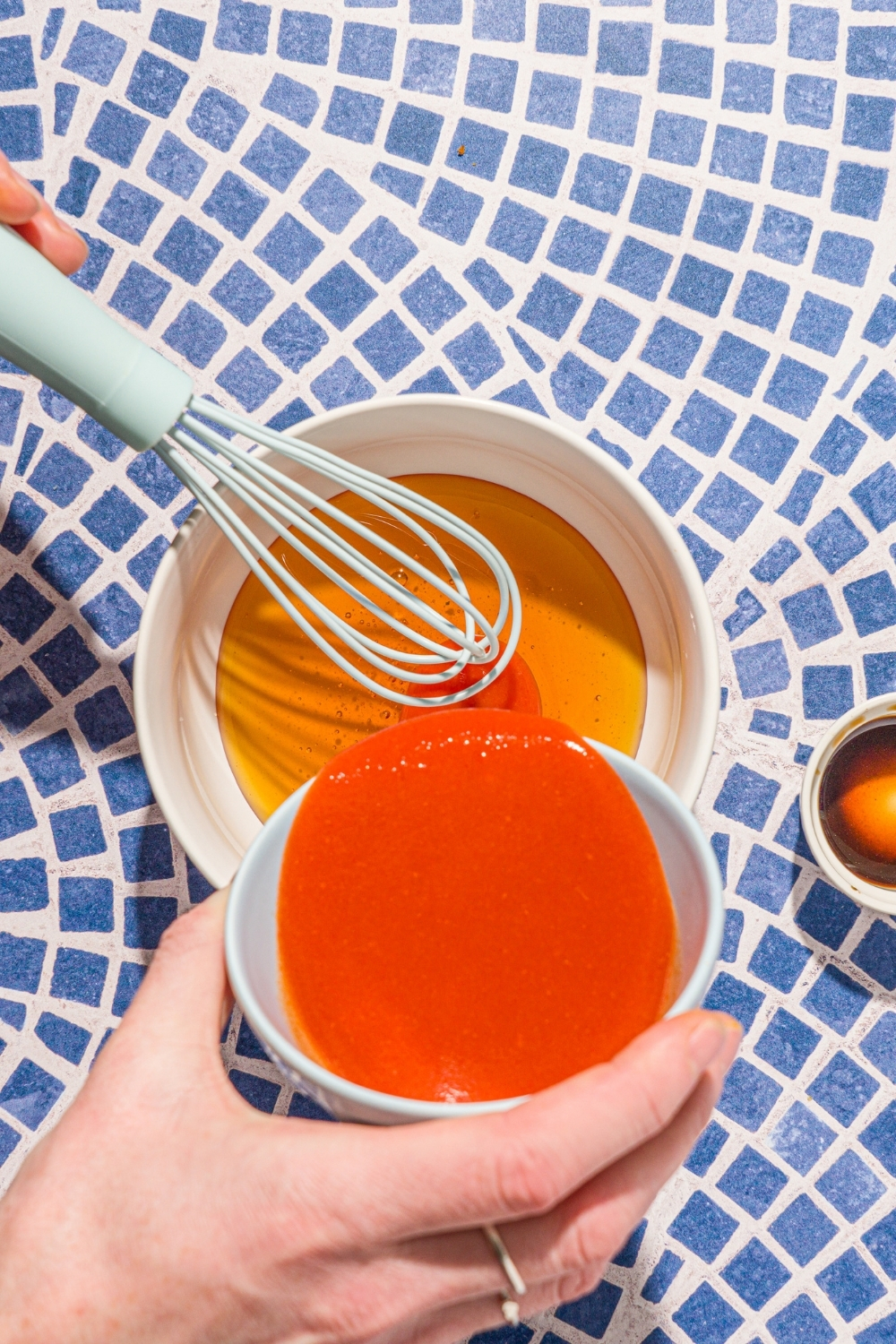 A bowl of hot sauce being poured into a bowl of honey with a whisk mixing the sauces. The bowl is on a blue tiled counter.