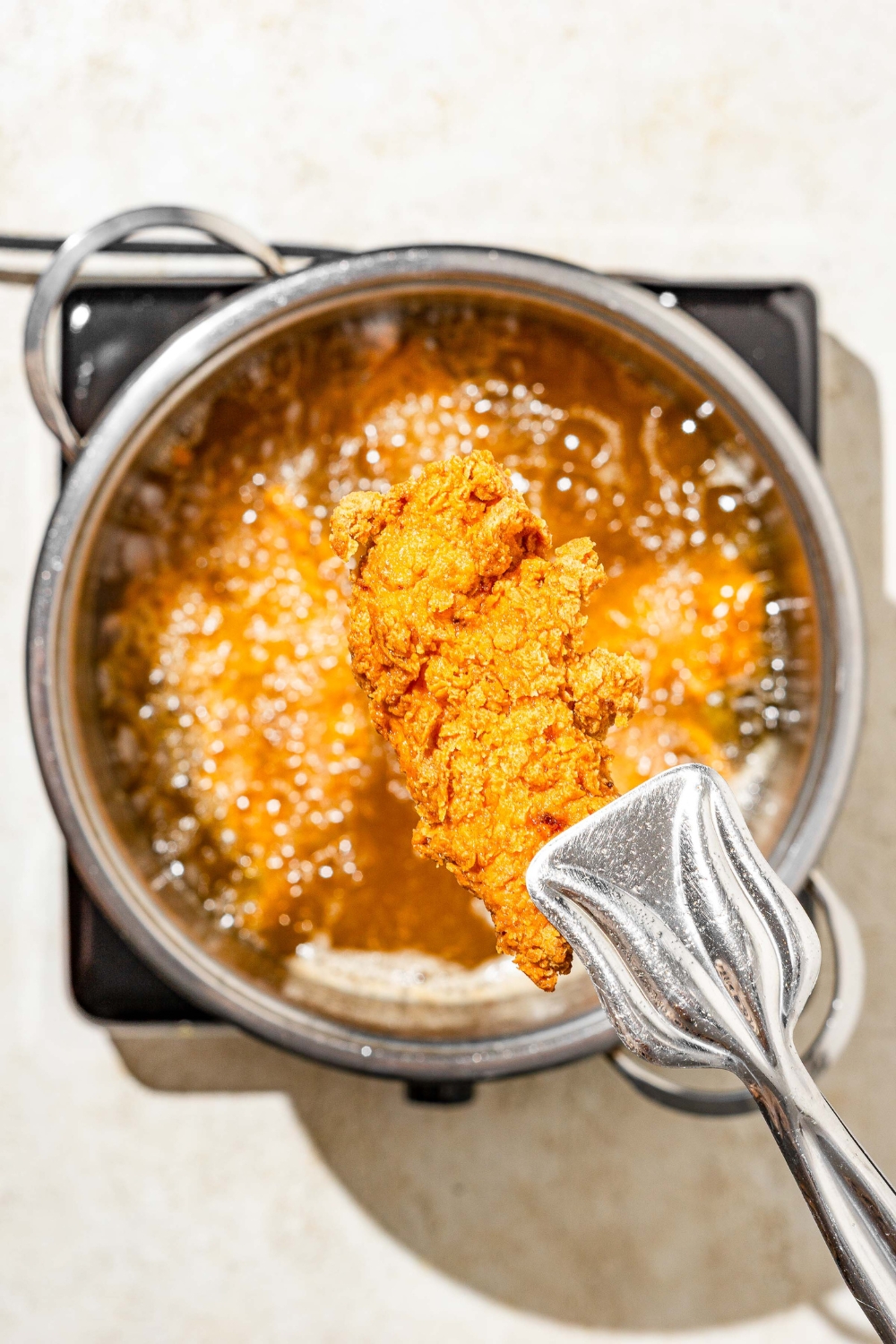 A close up of tongs holding a crispy fried chicken tender. There is a pot with breaded chicken tenders frying in oil blurred in the background.
