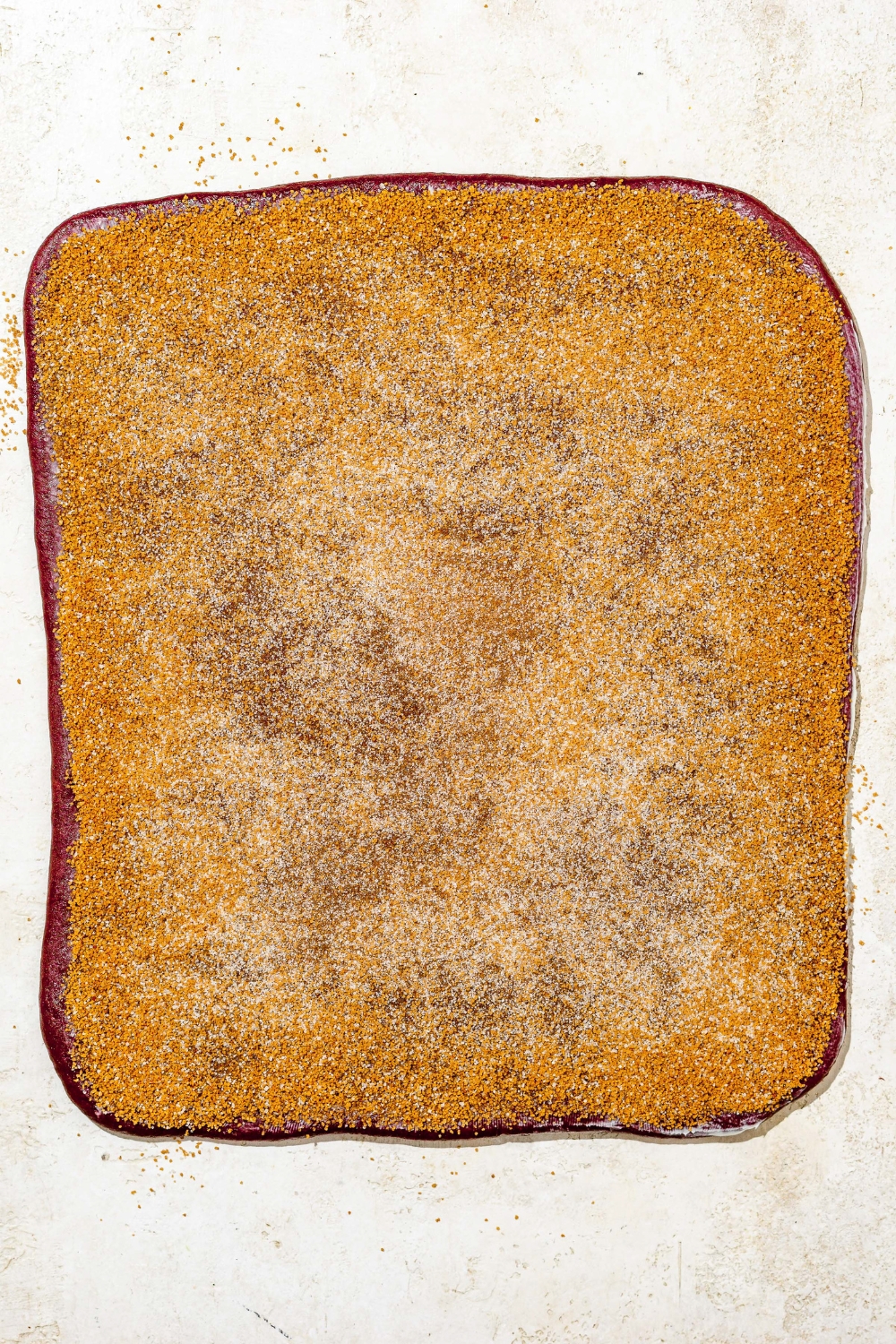 Red velvet cinnamon roll dough rolled out into a rectangle topped with cinnamon sugar mixture. The dough is on a tan counter.