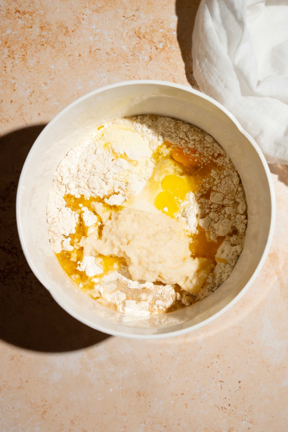 A white mixing bowl with ingredients to make dinner roll dough including flour, egg, milk, yeast, and seasonings. The bowl is on a tan counter with a white cloth napkin.